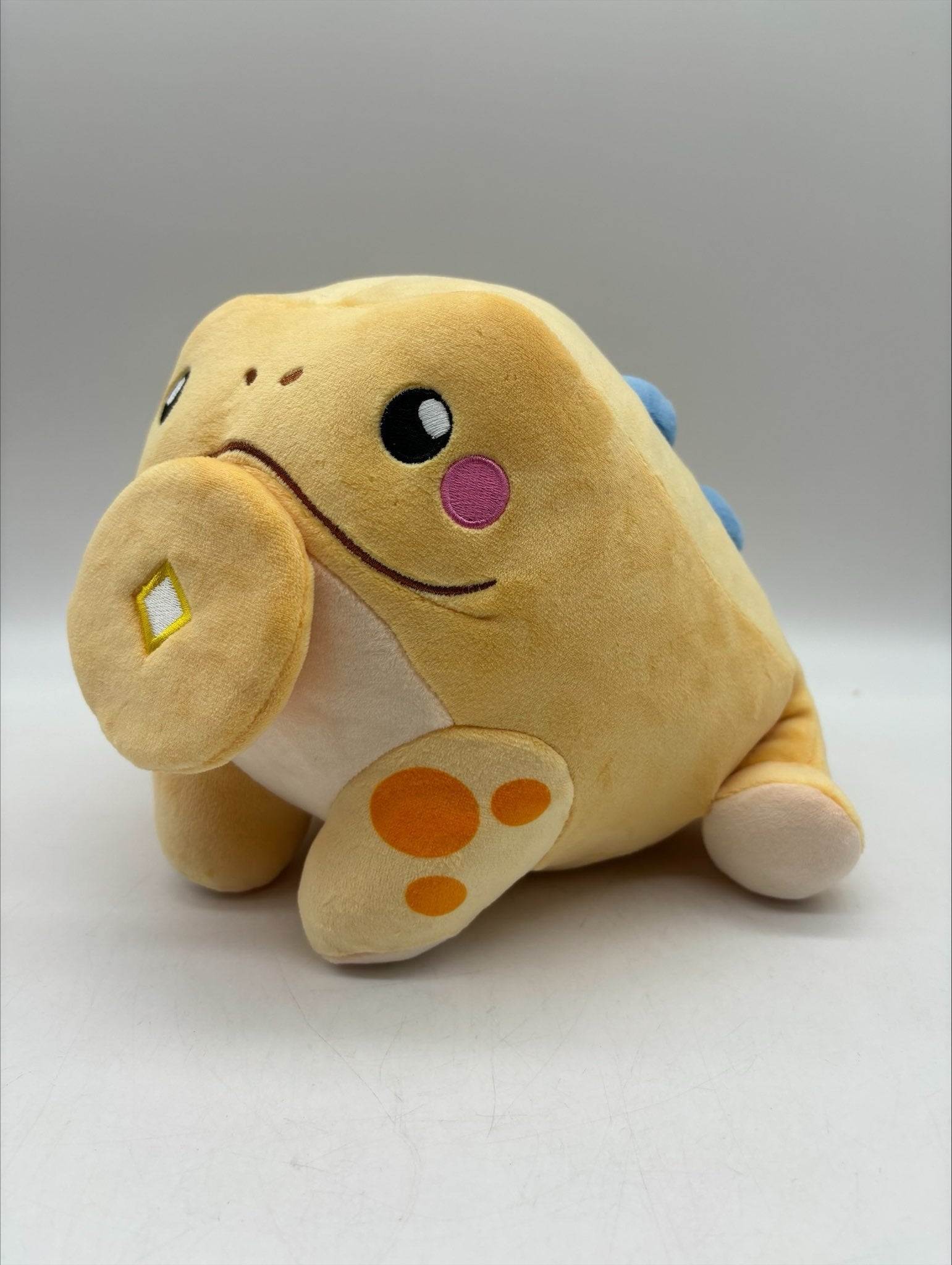 Lucky the Jin Chan plush by TOTYM, Chinese money frog from folklore, yellow‑orange with blue spots, holding a coin in its mouth, facing a left angle on a clean white background.