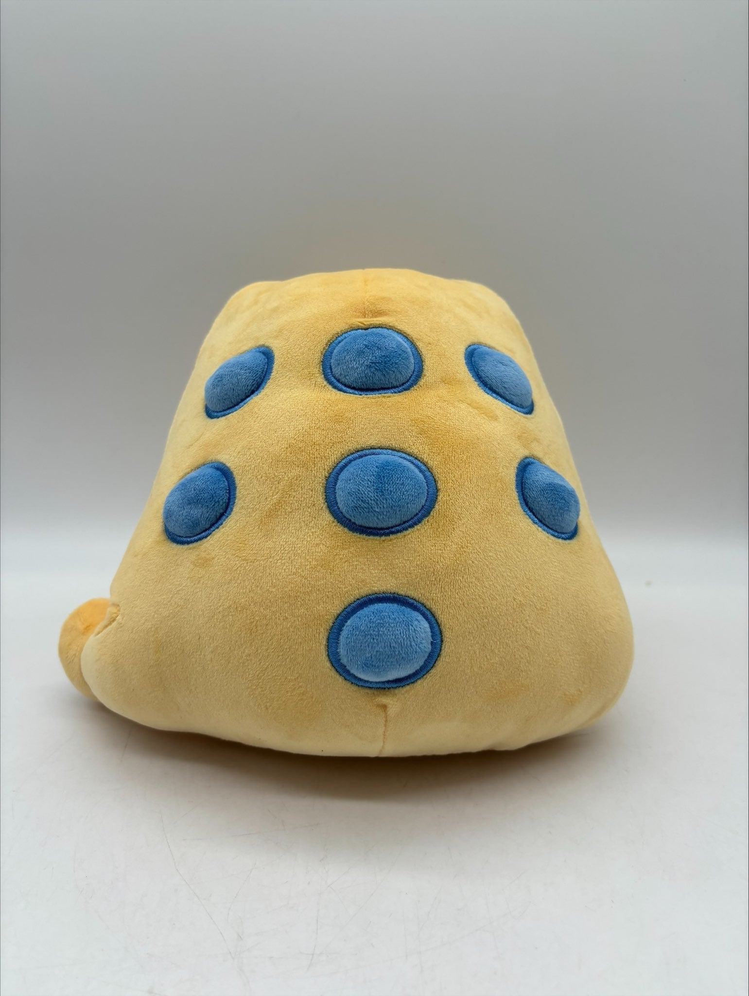 Lucky the Jin Chan plush by TOTYM, a Chinese money frog from folklore. Yellow‑orange body with blue spots, holding a coin in its mouth, shown from the back, centered against a clean white background.