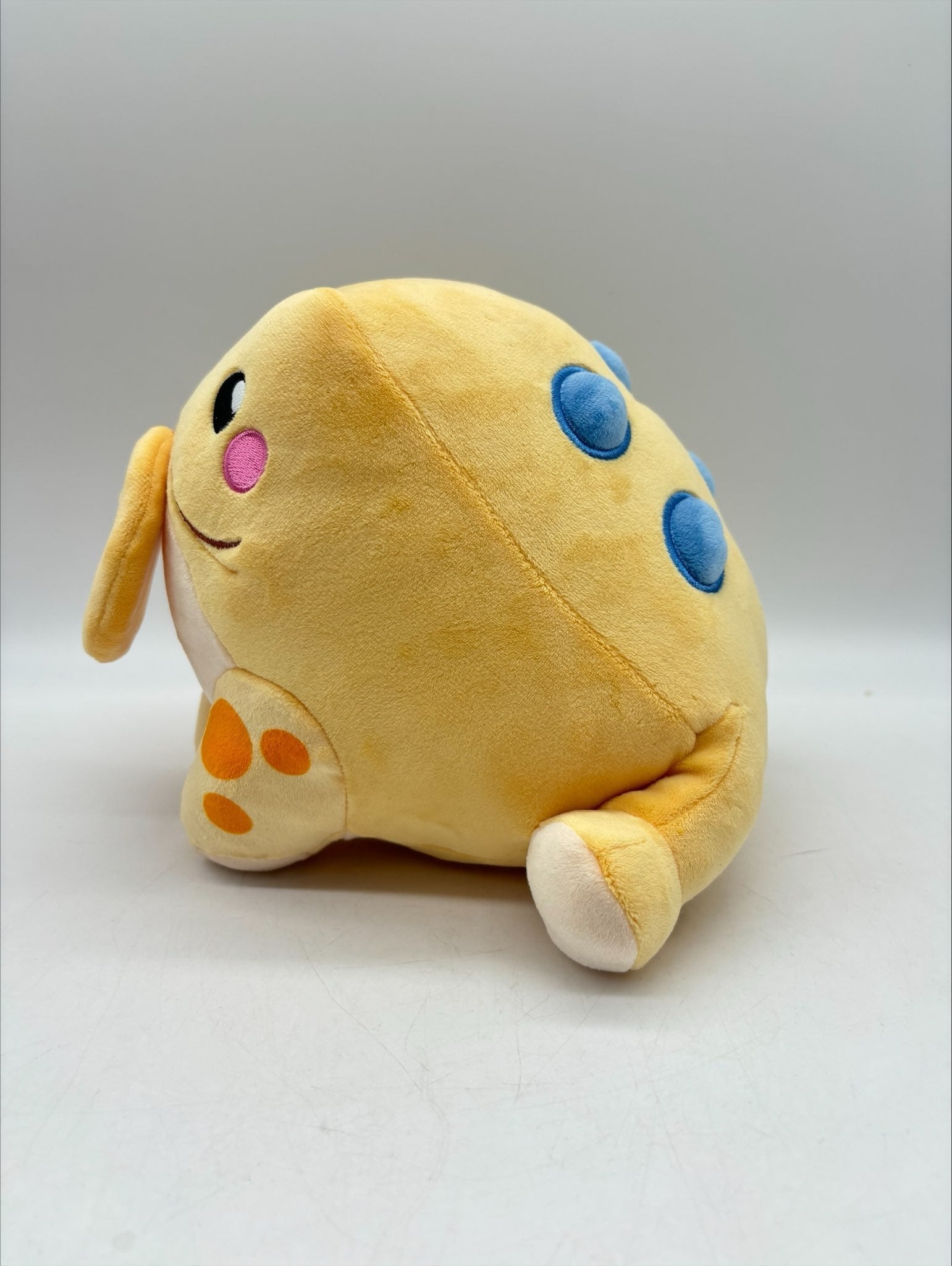 Lucky the Jin Chan plush by TOTYM, Chinese money frog from folklore, yellow‑orange with blue spots, holding a coin in its mouth, facing left on a clean white background.