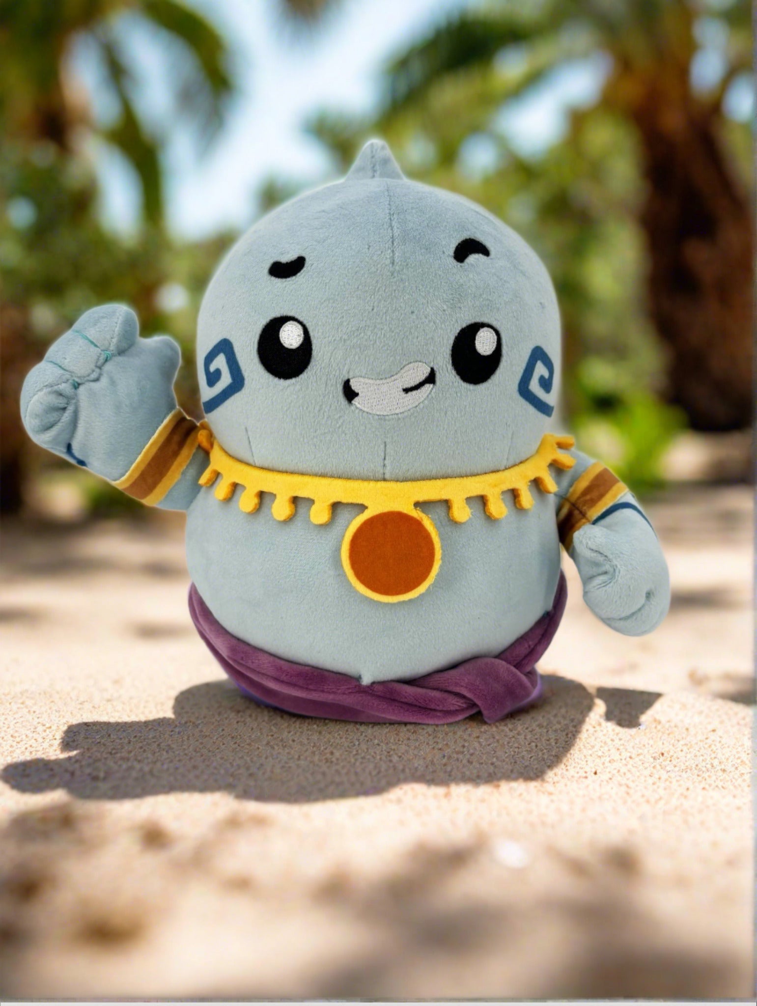 TOTYM's Djinn plush, a cute blue genie with golden accents and a purple sash, waving while standing on sand with palm trees in the background. Kawaii mythical creature plush perfect for collectors.