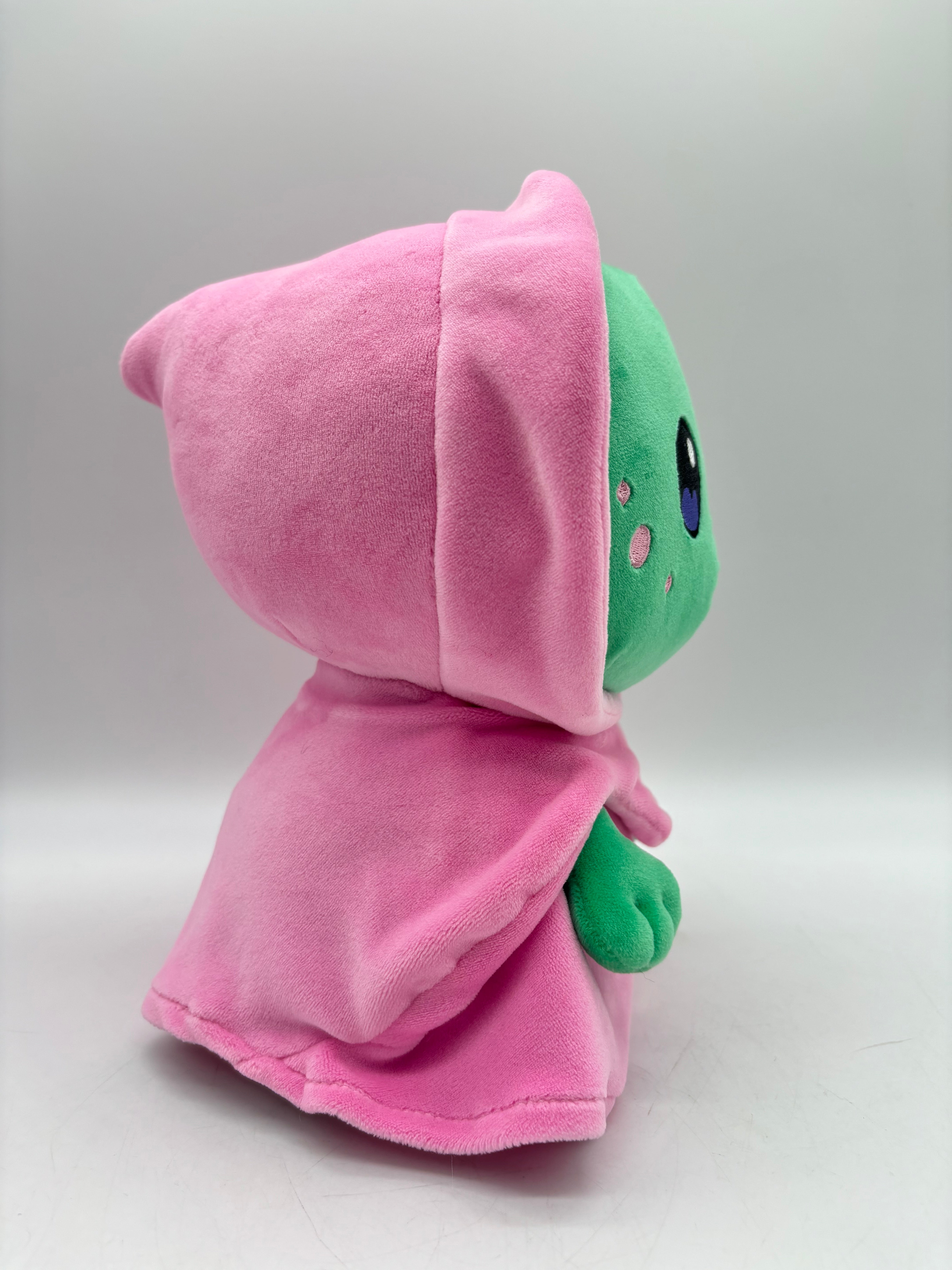 Side profile of TOTYM's Dreamwalker plush, showing its green face and pink hooded cloak. This charming, mystical plush toy is designed for fans of kawaii and collectible creatures.